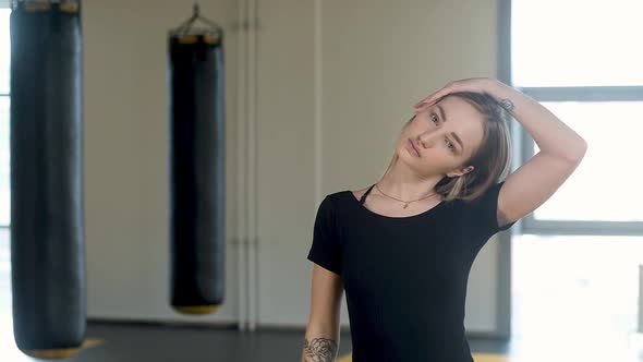 girl stretches her neck and shoulders against a backdrop of punching bags. alt