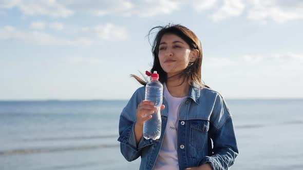 Woman Drinking Clear Mineral Water From Bottle at the Sea and Looking at Camera at the End. Health