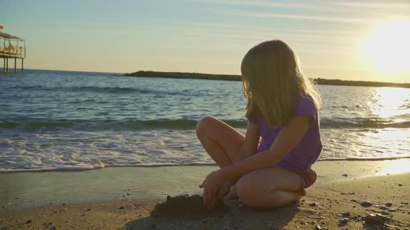 a Cute Little Girl Sits on the Seashore Playing in the Sand and with Pebbles alt