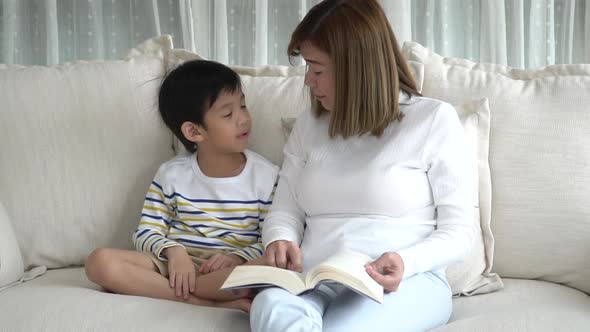Asian Mother And Her Son Reading A Book On Sofa In Living Room Slow Motion alt