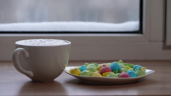 A Large Mug of Coffee with Milk or Mochacino and Colorful Sweets in a Plate on the Windowsill in