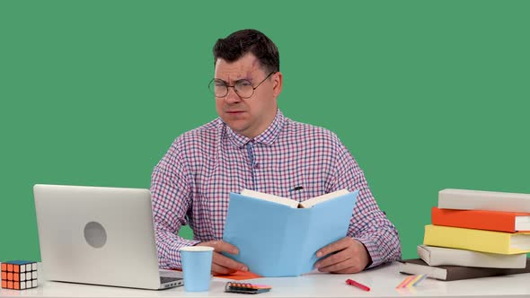 A Man with Glasses Sits at a Desk in Front of a Laptop and Reads a Book Nods His Head Negatively is alt