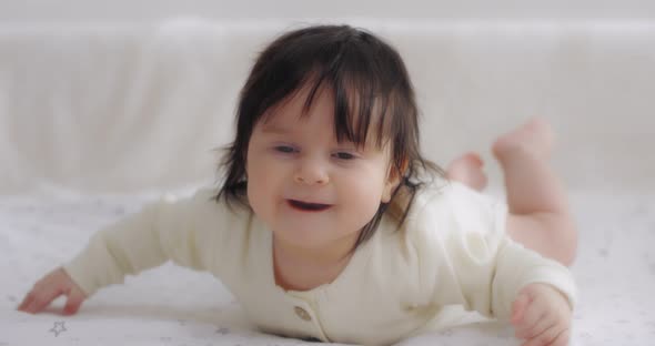 Funny Baby with Dark Hair Lies on Her Stomach on the Bed and Laughs