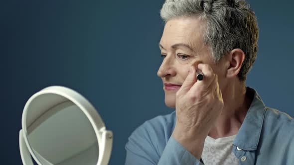 Wellgroomed Elderly Woman Applies a Cosmetic Product on Her Face with a Makeup Brush Sitting at Home