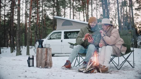Couple of Travelers Sits By a Campfire Looking at Phone in the Woods in Winter