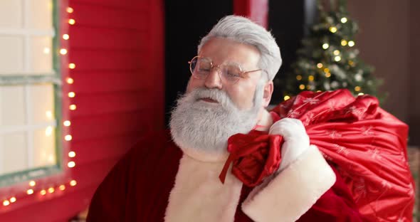 Sympathetic Santa Claus with a Large Sack of Gifts Walks Up to the New Year's Decorated Red House