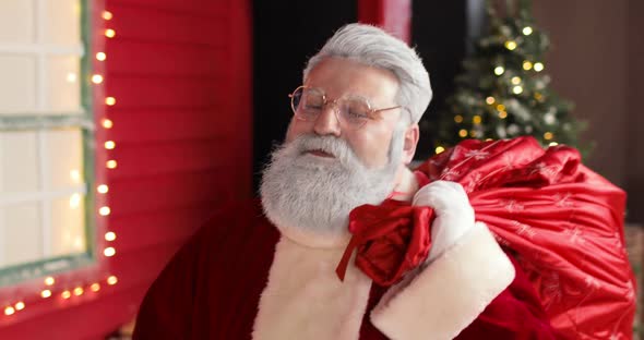 Sympathetic Santa Claus with a Large Sack of Gifts Walks Up to the New Year's Decorated Red House alt