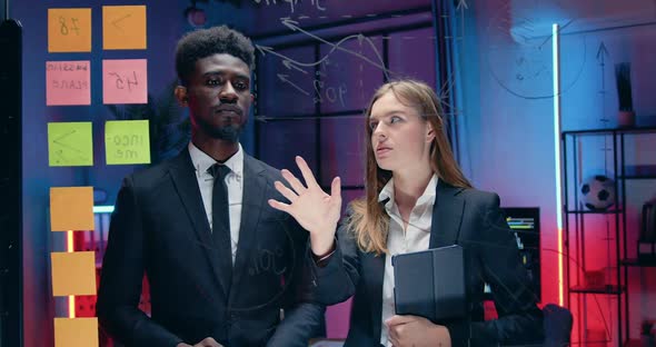 Male and Female Partners Discussing Joint Business Strategy Near Glass Wall in Evening Office alt