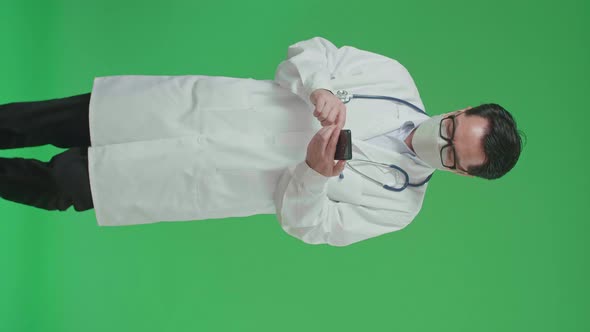 Asian Doctor With Stethoscope Wearing Mask, Using Mobile Phone While Walking On Green Screen alt