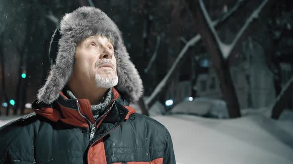 An Elderly Man with a Gray Beard in a Winter Hat is Standing on the Street and Enjoying the Snowy