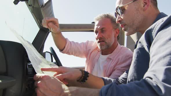 Happy caucasian gay male couple sitting in car reading map and pointing on sunny day at the beach alt