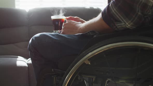 Midsection of caucasian disabled man in wheelchair holding cup of coffee in living room alt