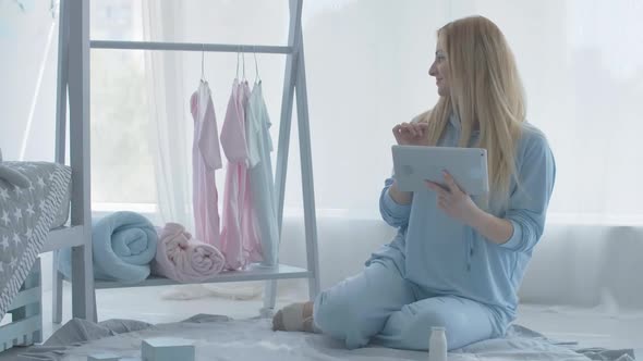 Smiling Pregnant Woman Using Tablet and Children Clothes Appear on Hanger. Portrait of Happy alt