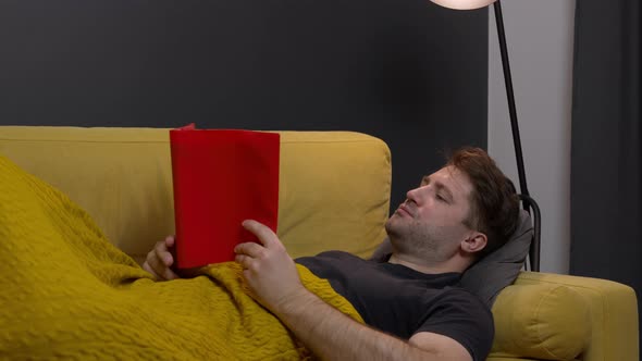 Young Unshaven Attractive Guy Lies at Home on Cozy Sofa and Reads Book