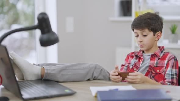 Relaxed Middle Eastern Little Boy Sitting with Feet on Table and Messaging Online on Smartphone alt
