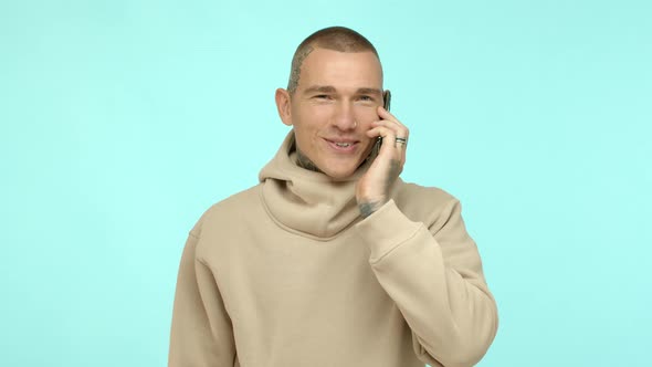 Cool and Stylish Tattooed Guy in Beige Hoodie Talking on Mobile Phone Smiling and Looking Happy