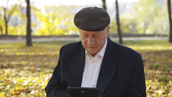 Senior Man in Cap Using Tablet and Telling on Sunny Autumn Background alt