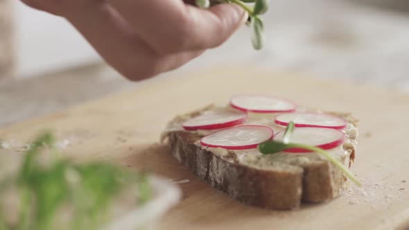 Man Cooking In The Kitchen. Man With Microgreens In Hand Sprinkles A Radish Sandwich. Vegan Sandwich alt