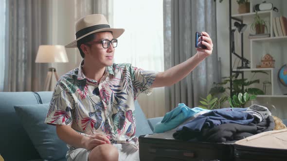 Asian Young Man Try Hat And Selfie While Packing Clothes In Suitcase At Home, Preparing For Vacation alt