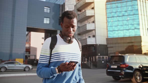 A Young Black Student in Stylish Clothes Sits and Talks on a Smartphone in the City