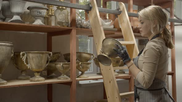 Young Woman Decorator Takes Out a Vase From a Shelf in a Workshop