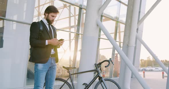 Man Standing Near Glass Wall with His Bike and Using Phone alt
