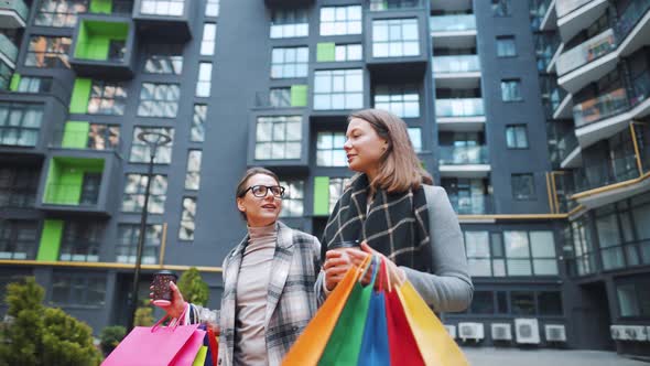 Two Happy Women Walk with Shopping Bags and Takeaway Coffee After a Successful Shopping and Talk alt