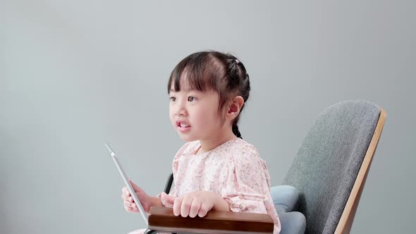 Asian child with fringe in hair sitting on chair laughing while using digital tablet. alt