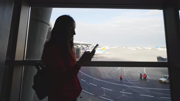 Businesswoman at Airport with Phone in Hands Against the Large Panoramic Window with Parking alt