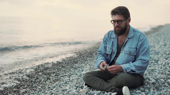 Bearded Man Is Throwing Stones Into Water of Sea, Sitting in Shore in Evening alt