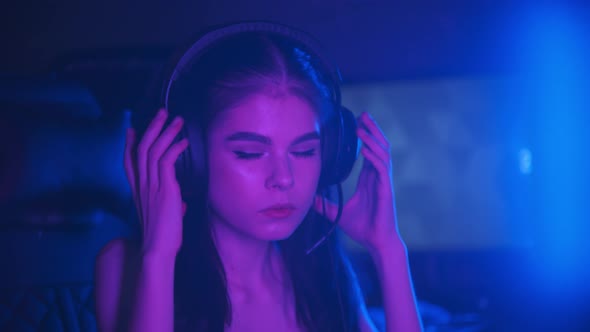 Young Attractive Woman with Light Makeup Sitting in Neon Gaming Club - Puts on Her Headphones and alt