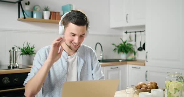 Man Puts on Headphones and Talks Online By Video Call Using Laptop in Kitchen