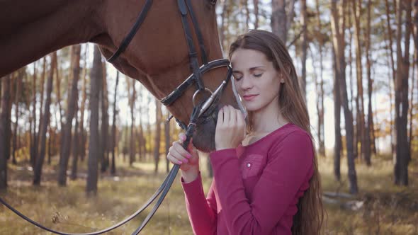 Close-up of a Young Caucasian Girl in Pink Clothes Caressing Horse in the Autumn Forest and Smiling alt