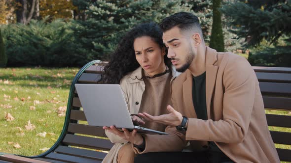 Worried Hispanic Young Couple with Laptop Sit on Bench in Autumn Park Unsuccessful Online Shopping alt