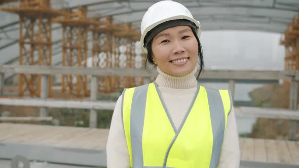 Portrait of Cheerful Asial Lady Wearing Helmet and Protective Vest Smiling in Building Area alt