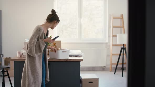 Young caucasian woman using mobile phone while unpacking stuff in new house. alt