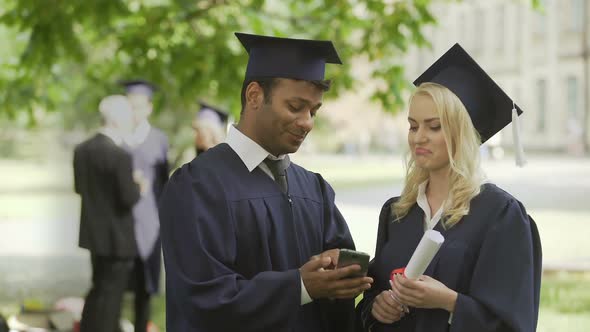 Young People in Graduate Uniform Looking at Phone Screen, Having Discussion, Job alt
