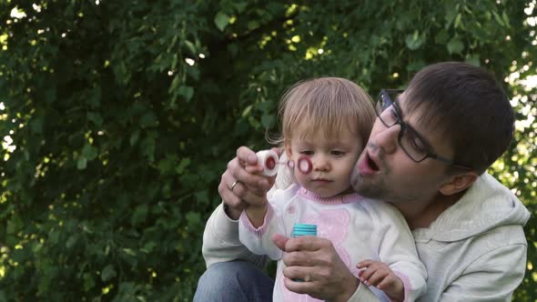 Father and Little Baby Girl Blow Soap Bubbles in Summer Park