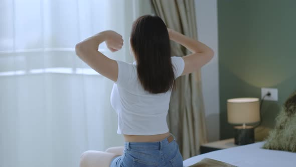 Back view of young woman raising her hands up and stretching arms sitting on the bed alt