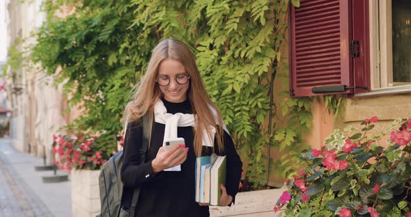 Young Blond in Casual Clothes Walking Near Buildings with Green Plants and Using Her Smartphone alt