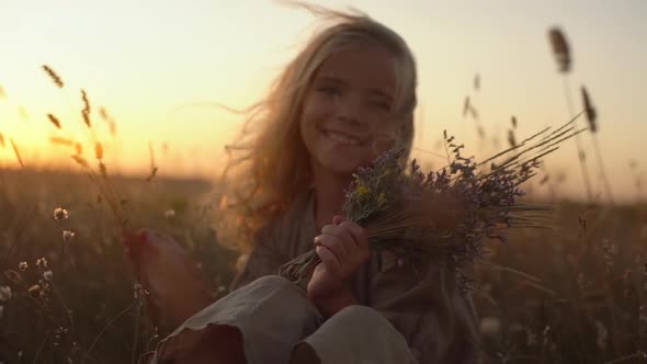 Portrait of Beautiful Little Girl with Blond Long Hair Sitting in Field Covered with Plants Holding alt