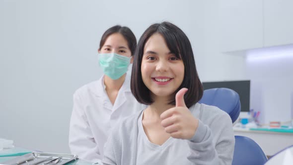 Asian dentist or doctor wearing mask sit behind young beautiful girl patient toothy smile in clinic. alt