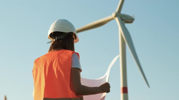 An Engineer of Wind Turbines Holding a Paper Project and Looking at Windmill in the Field alt
