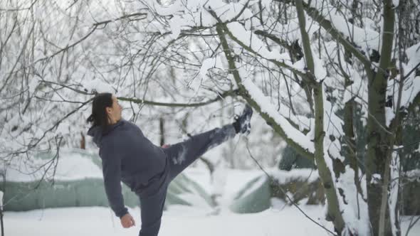 A Man Athlete with Long Black Hair Kicks a Tree Snow is Falling on Him alt