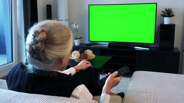 Old Caucasian Woman Watches Television in Living Room and Switches Program alt