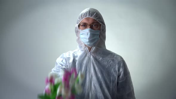 A Man in a Protective Suit, Medical Mask and Glasses Shows Flowers on a White Background