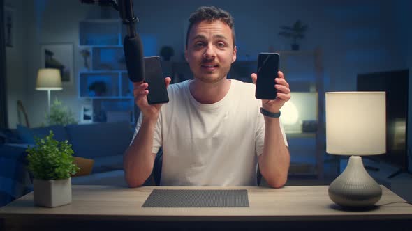 Young Handsome Man Makes an Overview of Two Smartphones at Home in the Living Room alt