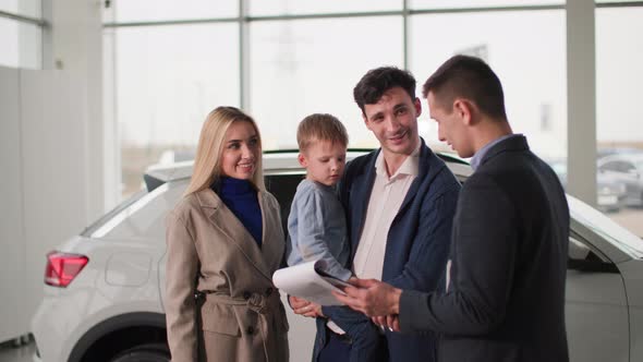 Family in Auto Show Happy Parents with Male Child Talking to Car Salesman About New Automobile Model