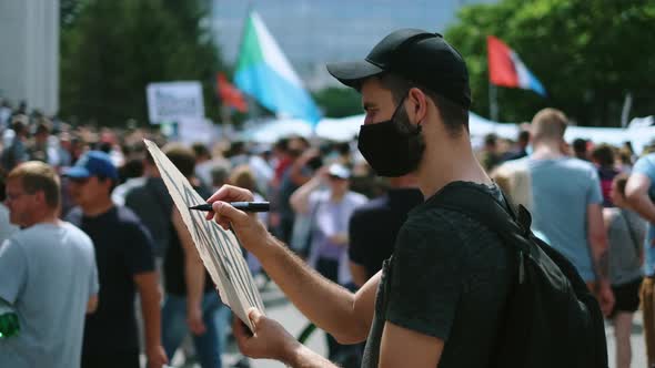 Male Protester Drawing Banner Sign in Hands with Marker in Rally Riot Crowd alt