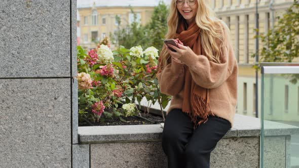Young Girl Student Sitting Outdoors in City with Colorful Bags Uses Smartphone for Shopping Online alt
